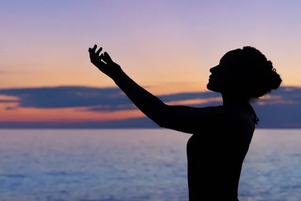 Silhouette of a woman with outstretched arms against a colorful sunset over the ocean.