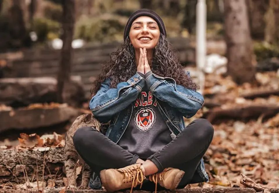 A woman with curly hair sitting cross-legged in a forest, wearing a denim jacket and a beanie, smiling with her hands in a prayer position.